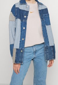 Jeansjacka med ett lapptäckedesign i olika nyanser av blått och grått. Har silverfärgade knappar och en bröstficka. Bärs över en ljusrosa topp.