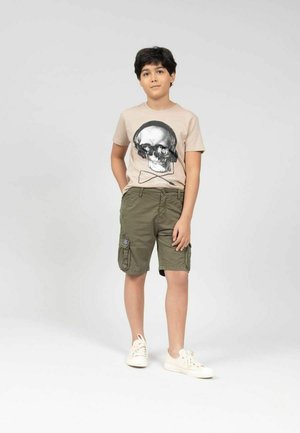 T-shirt beige avec un motif de crâne, associé à un short cargo vert olive. L'ensemble est complété par des baskets blanches.
