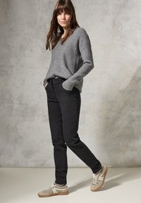 Suéter gris de canalé, jeans ajustados negros y zapatillas beige con detalles blancos. El modelo posa con las manos en los bolsillos frente a un fondo texturizado.