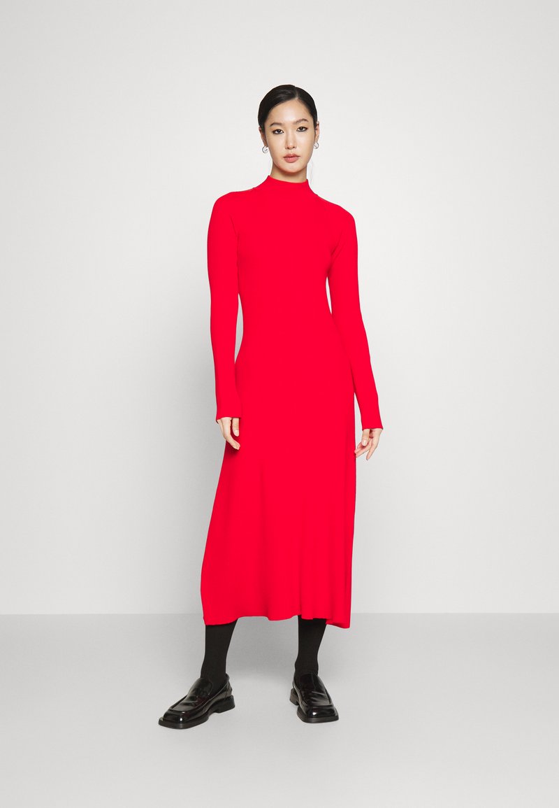Robe rouge à manches longues avec un col haut, texture lisse et silhouette fluide, associée à des mocassins noirs et des collants.