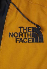 Logo The North Face brodé en noir sur tissu jaune moutarde avec un cordon noir et un bloqueur en plastique au-dessus du texte.