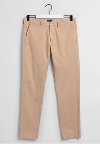 Chinos kaki avec devant plat, poches latérales et texture lisse, dotés d'une fermeture à bouton et d'une passants pour ceinture. Suspendus à un crochet noir.