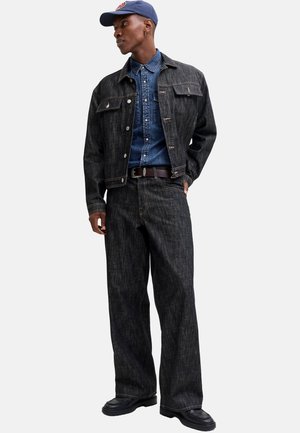 Man draagt een donker denim jack en broek, een blauw denim overhemd, een marineblauwe pet, zwarte schoenen en een bruine riem, staand met één hand in de zak.