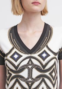 Tričko s výstřihem do V, které představuje geometrický vzor v černé, zlaté a modré barvě, s texturovanými akcenty a krátkými rukávy. Hladký povrch tkaniny.