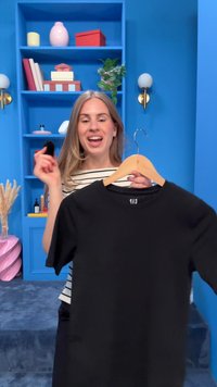 Femeie zâmbitoare într-o cameră albastră ține un tricou negru pe umeraș într-o mână și un obiect mic negru în cealaltă, stând în fața unei biblioteci.