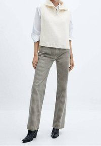 Cremefarbener, gestrickter ärmelloser Pullover mit hohem Kragen, kombiniert mit grauen, gestreiften, weit geschnittenen Hosen und schwarzen Stiefeletten mit Absatz.