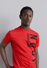 Punane puuvillane T-särk, millel on julged mustad tätoveeringud "DSQUARED2 IRON" külje peal. Sobiv disain ümmarguse kaeluse ja lühikeste varrukatega.