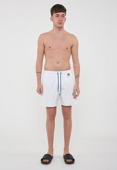 Shorts de bain blancs avec cordon bleu, et poches latérales. Le modèle est pieds nus, portant des sandales noires, contre un fond neutre.
