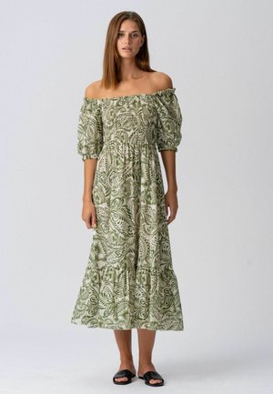 Fiatal nő, váll nélküli zöld-fehér mintás midi ruhában, puffos ujjakkal és fekete papucs szandálban, egyszerű háttér előtt állva.