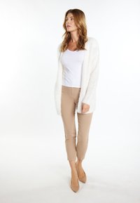 Cardigan alb pufo peste un top alb din bumbac, asociat cu pantaloni bej mulați și pantofi cu toc nude, stând pe un fundal deschis.