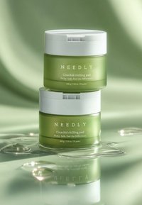 Vihreitä lasipurkkeja valkoisilla kansilla, joiden etiketeissä lukee "NEEDLY Cicachid Chilling Pad", pinottuna ja heijastuen vesipinnassa, sileällä, minimalistisella muotoilulla.
