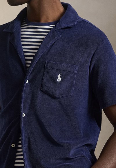 Μπλε ναυτικό κοντομάνικο πουκάμισο από υφασμάτινη υφή, με τσέπη στο στήθος που φέρει κεντημένο λογότυπο, φορεμένο πάνω από ριγέ μπλε ναυτικό και λευκό τοπ.