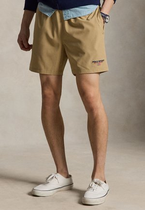 Vyras, dėvintis smėlio spalvos Polo Sport šortus, šviesiai mėlyną marškinius, tamsiai mėlyną megztinį, baltus sportinius batelius ir laikrodį ant riešo, stovintis su rankomis kišenėse.