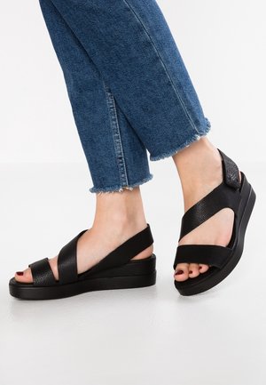 Sandales compensées noires avec des brides en cuir croisées, présentant une finition texturée. Semelle souple et design à bout ouvert. Portées avec un jean court en denim.