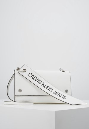 Sac d'épaule blanc Calvin Klein Jeans avec une sangle logo audacieuse posé sur un socle blanc sur un fond uni.