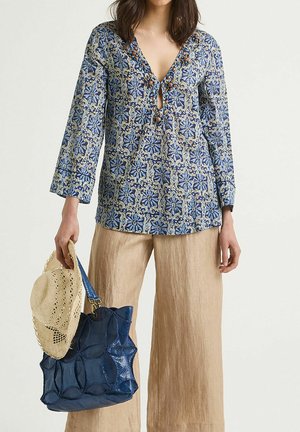 Blaue gemusterte Bluse mit einem Schlüsselloch-Ausschnitt, langen Ärmeln und dekorativen Perlen; kombiniert mit beigen Leinenhosen, einem Strohhut und einer blauen Tragetasche.