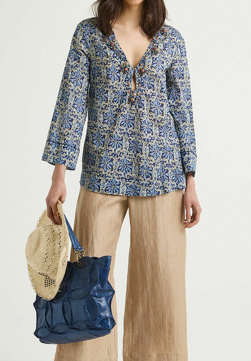 Blusa blu a fantasia con scollatura a occhiello, maniche lunghe e perline decorative; abbinata a pantaloni di lino beige, un cappello di paglia e una borsa a tracolla blu.