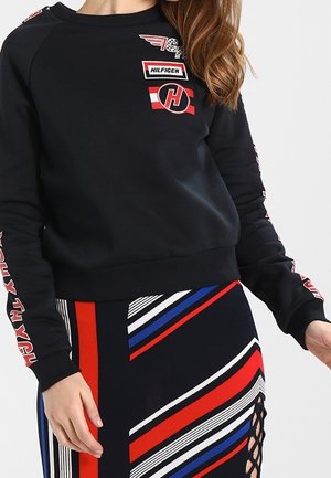 Kobieta w czarnej bluzie z czerwonymi i białymi nadrukami oraz spódnicy z czerwonymi, białymi, niebieskimi i czarnymi ukośnymi paskami oraz bocznym sznurowaniem.