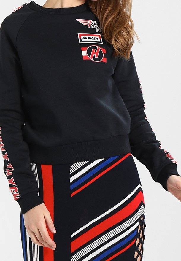 Femme portant un sweat-shirt noir avec des logos rouges et blancs, et une jupe présentant des rayures diagonales rouges, blanches, bleues et noires avec un détail lacé sur le côté.