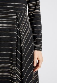Robe noire avec un motif rayé beige. Manches longues et design plissé. Le tissu semble doux avec une légère texture. Main partiellement visible.