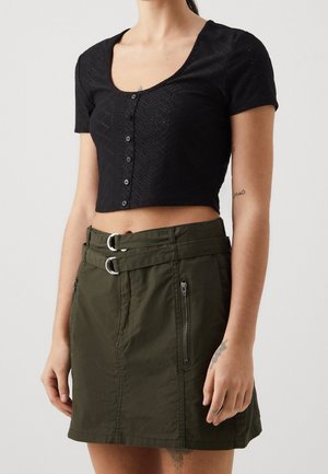 Top court noir avec un motif texturé et une fermeture à boutons, associé à une jupe vert olive avec des accents à double boucle et des fermetures éclair sur le côté.