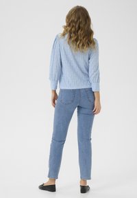 Lichte blauwe, gemarmerde top met een ruitpatroon, 3/4 mouwen; gecombineerd met aansluitende, middelhoge jeans en zwarte lage schoenen.