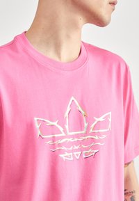 Pink cotton t-shirt with a round neck, featuring a shiny silver graphic logo on the front. The fabric has a smooth, soft texture.