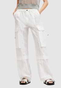 Een persoon in ruime witte cargobroek met elastische boorden en grote zijzakken, gecombineerd met open teen zilveren sandalen en een grijs mesh-top.
