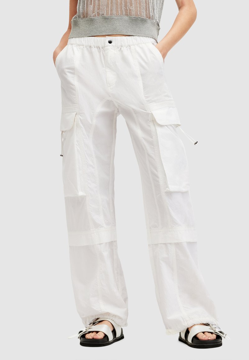 Een persoon in ruime witte cargobroek met elastische boorden en grote zijzakken, gecombineerd met open teen zilveren sandalen en een grijs mesh-top.