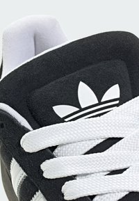 Čierna semišová teniska s bielymi textúrovanými šnúrkami. Má ikonický detail so štyrmi pruhmi a na jazyku logo pripomínajúce kvet.