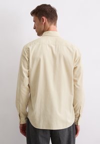 Lichtbeige lange mouw shirt van een gestructureerde stof, met een klassieke kraag, knoopmanchetten en een afgeronde zoom, gedragen door een model vanachter.