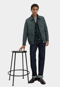 Uomo con giacca verde a zip, cardigan scuro, jeans e scarpe marroni, in piedi con una mano appoggiata su uno sgabello alto nero.