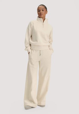 Vrouw staat met haar handen in haar zakken, draagt een crèmekleurige zip-up trui en brede broekspijpen met slippers op een effen achtergrond.