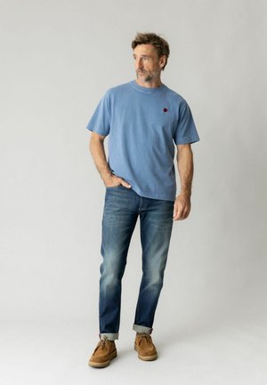Mand stående med hånden i lommen iført blå t-shirt, opkaldte jeans og brune sko mod en ensfarvet lys baggrund.