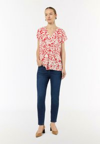 Rote und weiße gemusterte Bluse mit V-Ausschnitt, kurzen Ärmeln und lockerem Schnitt, kombiniert mit dunkelblauen Skinny-Jeans und beigen High Heels.