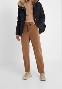 Sort dunjakke med hætte i kunstpels, beige rullekravesweater og brune fløjlsbukser. Beige chunky sneakers fuldender outfittet.