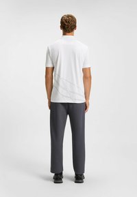 Weißes Poloshirt mit kurzen Ärmeln und grauen diagonalen Linien auf dem Rücken, kombiniert mit dunkelgrauen strukturierten Hosen und schwarzem Schuhwerk.