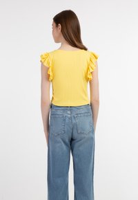 Gul ribbet top med volangærmer, parret med højtaljede blå denim jeans, vist fra rygsiden med detaljer om tøjet.