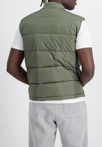 Olivengrüne Daunenjacke ohne Ärmel mit Stehkragen und gesteppter Optik,über einem weißen Hemd getragen, dazu sichtbare graue Jogginghose.