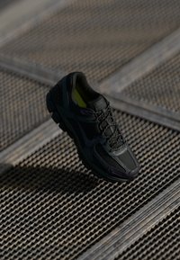 Scarpa sportiva nera con parte superiore in mesh e sintetico, superficie testurizzata, suola ammortizzata e lacci. Accenti interni gialli vivaci.