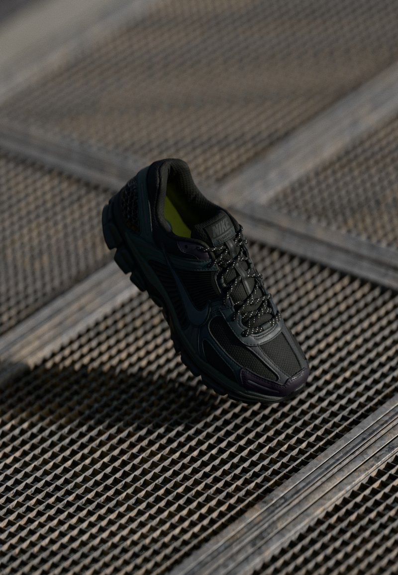 Scarpa sportiva nera con parte superiore in mesh e sintetico, superficie testurizzata, suola ammortizzata e lacci. Accenti interni gialli vivaci.