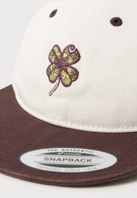 Casquette snapback blanche et marron avec un trèfle à quatre feuilles brodé en vert et marron sur le panneau avant et un autocollant de marque sur la visière.