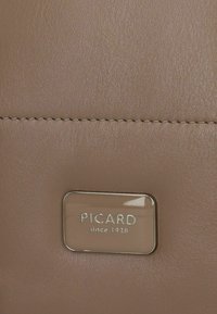 Taupe leather handbag featuring a shiny silver rectangular logo plate with "PICARD since 1928." Smooth texture and subtle stitch detailing.