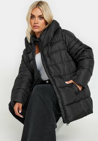 Svart puffjacka med huva, quiltat mönster, dragsko detaljer, dragkedjestängning och sidofickor, bärs över en vit topp och mörka byxor.