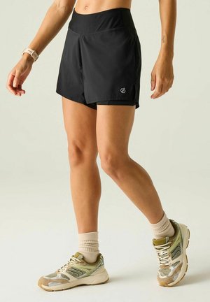 Vrouw in zwarte sportshort, beige sokken en beige hardloopschoenen staand met licht gebogen benen.