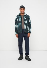 Fleece jacket in dark teal with white and burgundy patterns, paired with gray top and navy checkered pants. Black sneakers complete the outfit.