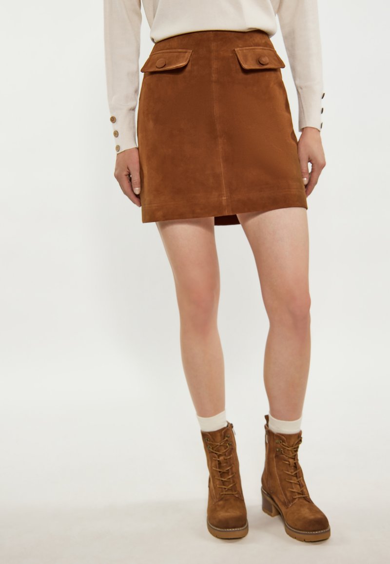 Mini-jupe en daim brun avec deux poches avant, design plat à l'avant, associée à des bottines brunes à lacets et des chaussettes blanches.