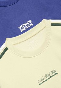 Two folded crewneck t-shirts, one blue with "VENICE BEACH" text and one light beige with green stripes and "New South Wales East Coast of Australia" text.