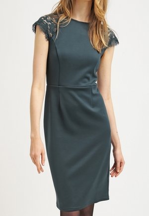 Femme portant une robe ajustée vert foncé jusqu'aux genoux avec des manches courtes en dentelle, debout devant un fond clair uni.