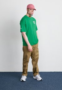 Grøn bomulds T-shirt med korte ærmer; khaki bukser; lyserød baseballkasket; hvide og sorte sneakers; ensfarvet baggrund.